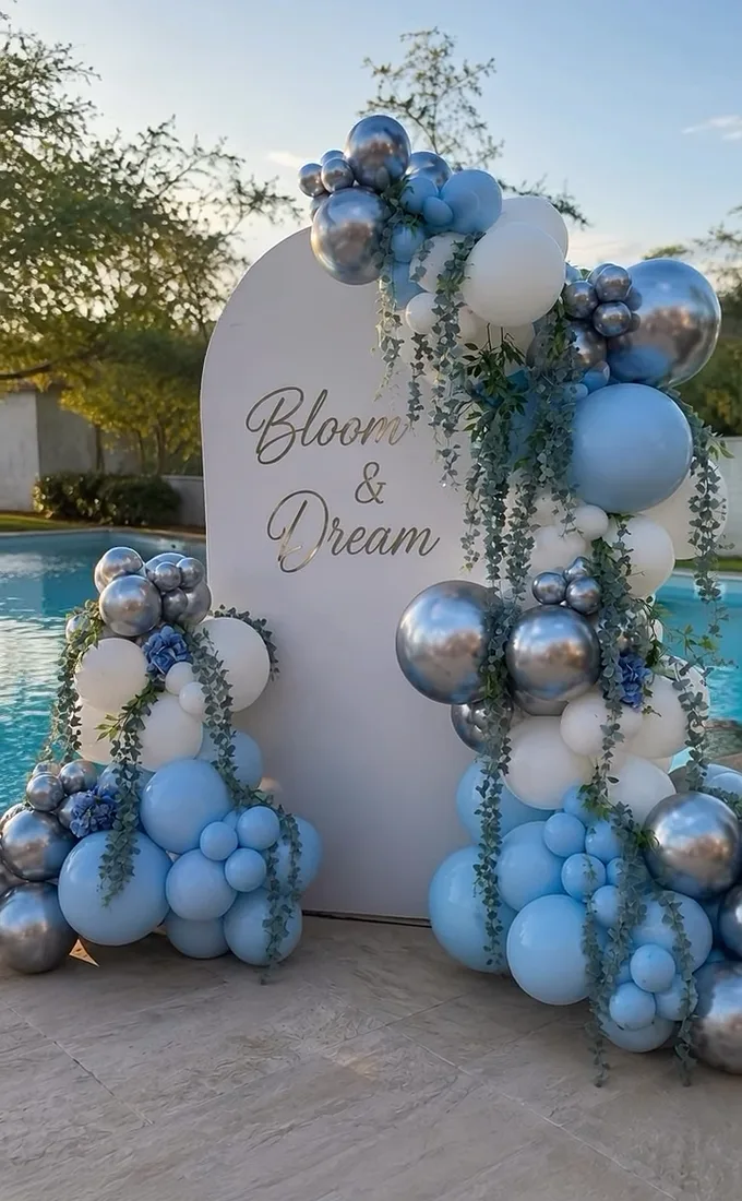 Backdrop blanco con el nombre Bloom and Dream, globos azules y plateados y follaje, junto a una alberca.