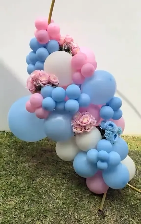 Aro dorado con globos rosas y azules y flores, montado en un jardín para una revelación de género.