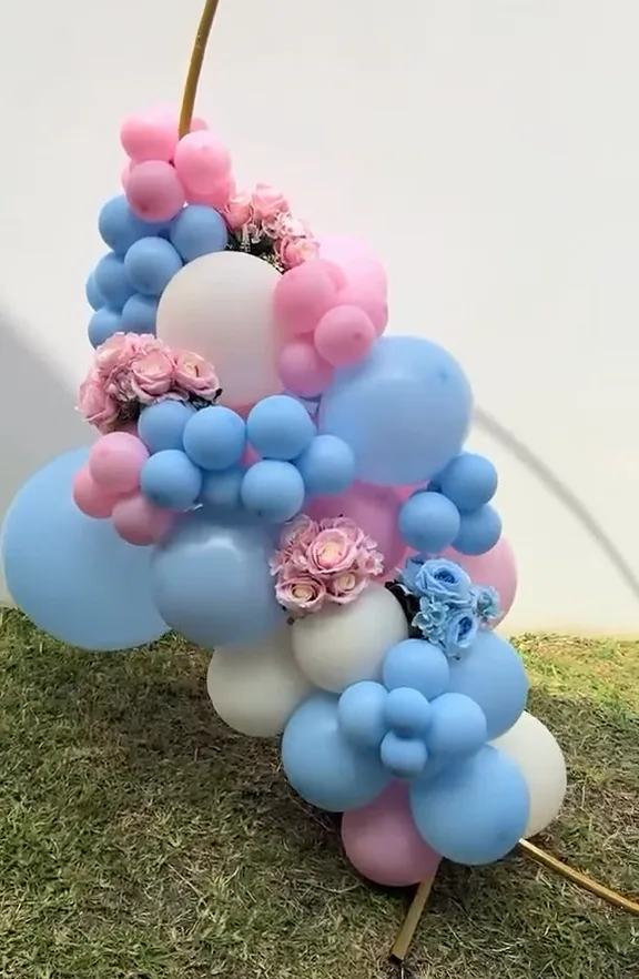 Racimo de globos rosas, azules y crema con flores sobre un aro dorado, en el césped.